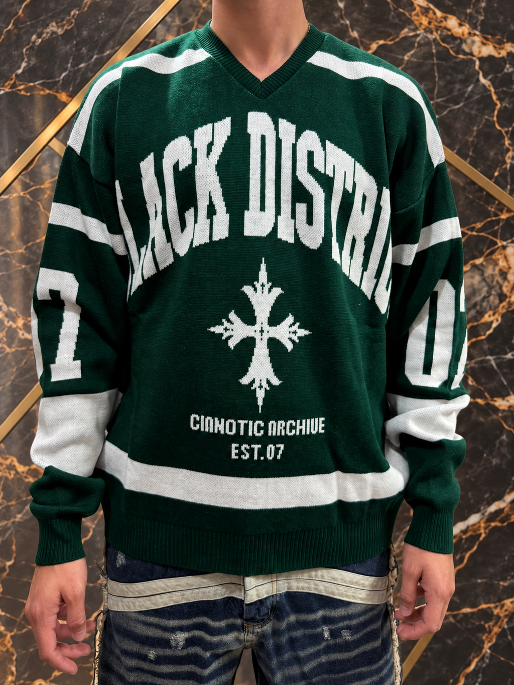 Maglione verde Black District indossato, collo a V e dettagli avorio su fondo nero e oro, senza volto
