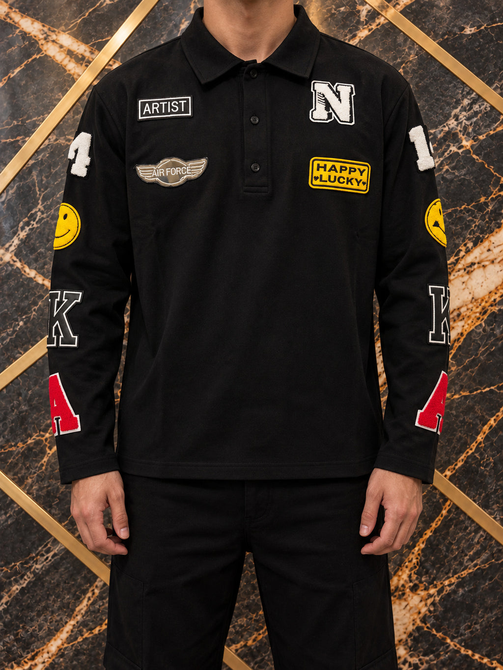 Polo nera con patch colorate, indossato, inquadratura senza volto su marmo nero e oro — immagine rielaborata