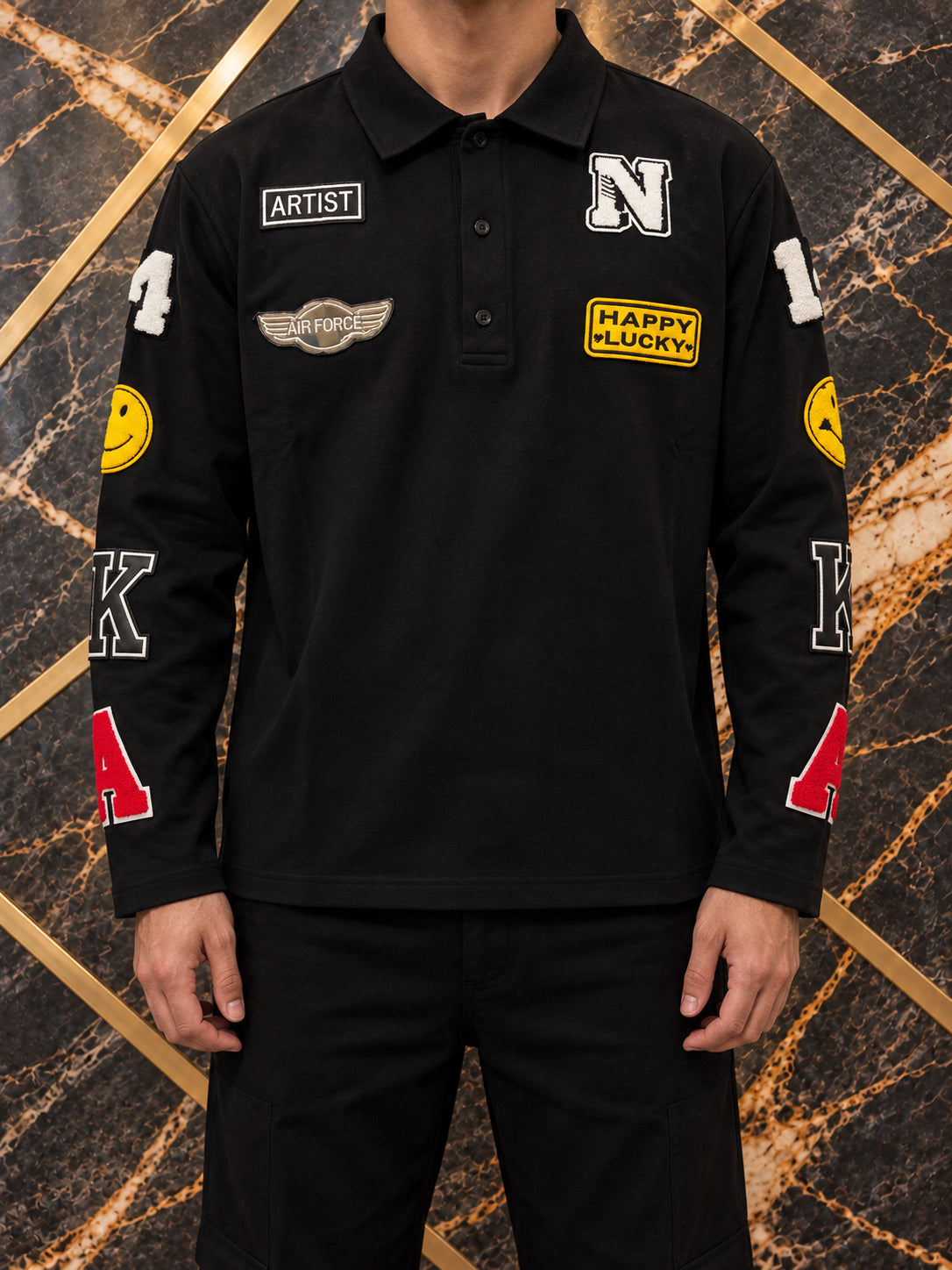Polo nera con patch colorate, indossato, inquadratura senza volto su marmo nero e oro — immagine rielaborata