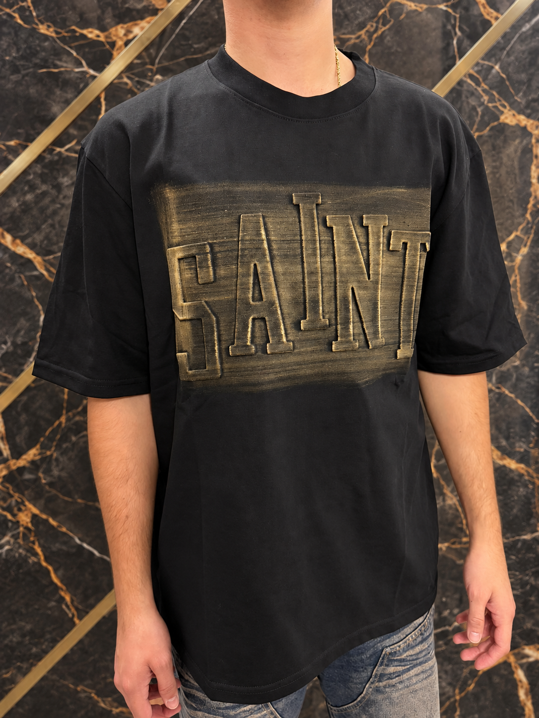 T-shirt nera SAINT indossata, scritta dorata e inquadratura senza volto su fondo nero e oro